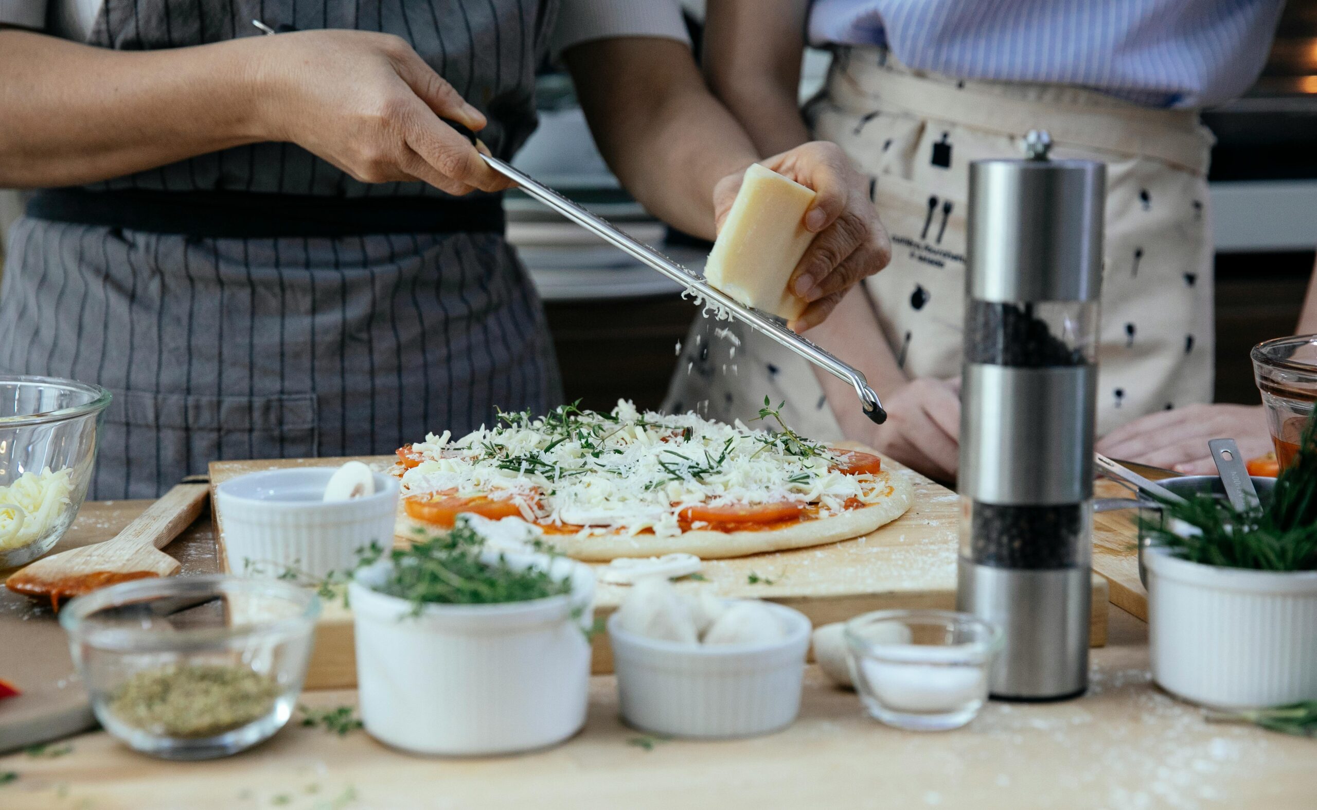 Unrecognizable female cooks grating cheese on homemade pizza at table with various ingredients and condiments in kitchen during cooking process